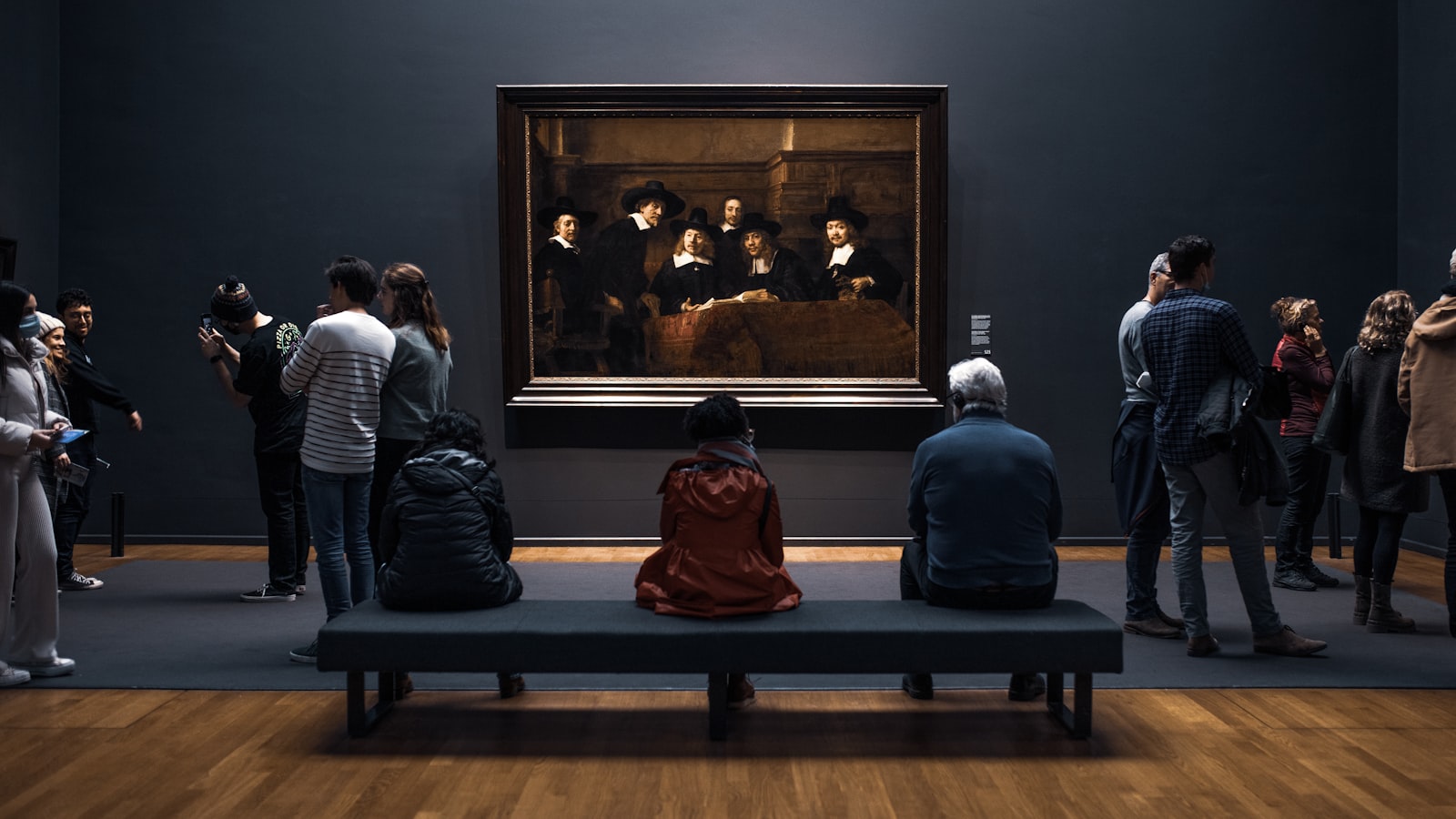 Museum visitors sitting and standing in a dark gallery while viewing a large framed painting